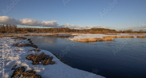 March sunny evening by the river. Blue sky over the horizon. A picturesque landscape, early spring, a river with snow-covered banks, dry grass and bushes. The first thaws, the snow is melting