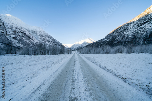 A straight dirt road in the mountains covered with white snow, but above the mountains there are beautiful clear blue skies