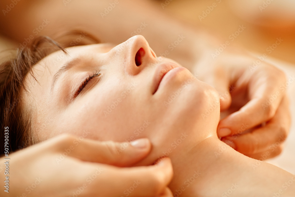 Warm toned closeup of young woman enjoying face massage session in SPA center with eyes closed