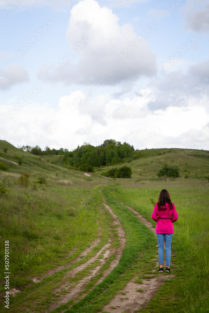 A woman in a pink raincoat looks at a mountain road. The concept of hiking in the mountains, trekking, outdoor recreation.