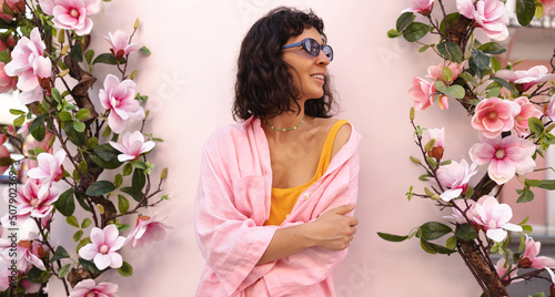 Pretty young caucasian woman averts her gaze posing on pink background with flowers. Brunette wears sunglasses and summertime shirt. Mood, lifestyle, concept . photo