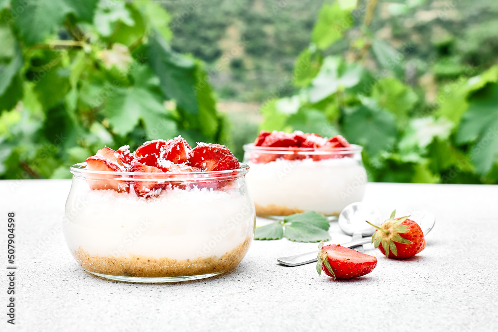 Strawberry desserts with fresh strawberries, cream cheese or natural yoghurt and oat granola in glass jars on light surface and nature background. Delicious healthy organic summer snack or breakfast