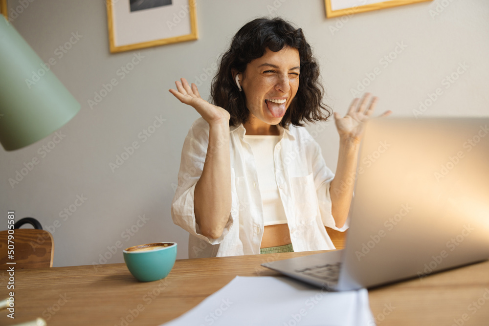 Funny young caucasian girl is sticking her tongue out on laptop camera sitting at her desk. Brunette wear white shirt is waving her arms and squinting. Concept of fun and technology Funny young caucasian girl is sticking her tongue out on laptop camera sitting at her desk. Brunette wear white shirt is waving her arms and squinting. Concept of fun and technology