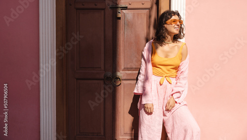 Beautiful young caucasian woman poses in sun outside city building. Brunette wears casual summer clothes and sunglasses. Lifestyle concept 