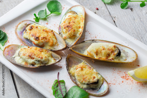 Fototapeta Naklejka Na Ścianę i Meble -  Mussels baked in cheese sauce on a rectangular platter. Decorated with lettuce leaves, spices and lemon wedges