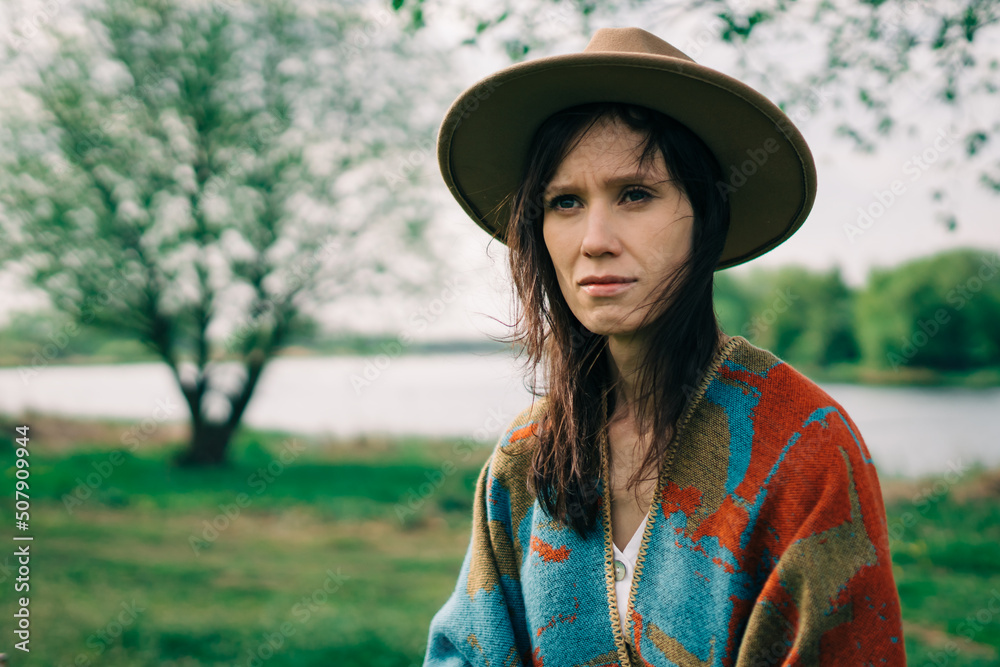 Portrait of a stylish woman in a hat and poncho at cloudy autumn day stands on countryside with river.