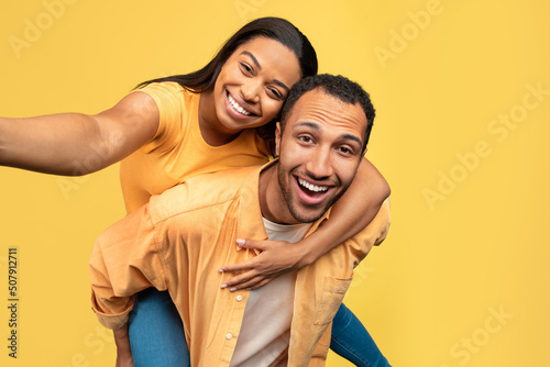 Cool young black guy giving piggyback ride to his girlfriend, making selfie together on yellow studio background