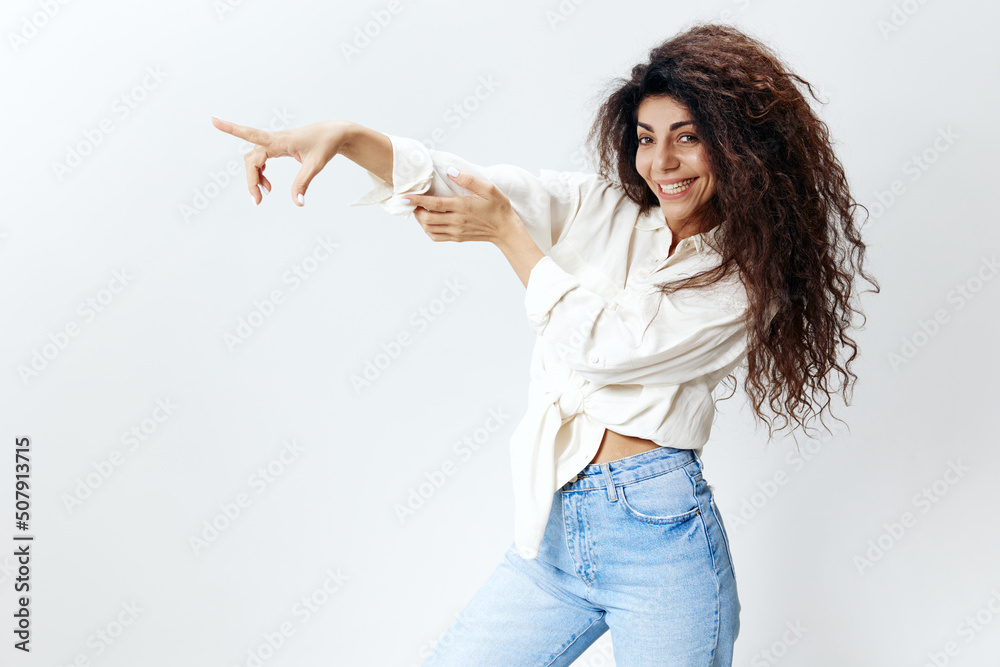 LOOK AT THIS. Portrait of happy cheerful pretty curly Latin woman staying near white studio wall background smiling, look at the left and point finger at free place, Copy space for clothes sales LOOK AT THIS. Portrait of happy cheerful pretty curly Latin woman staying near white studio wall background smiling, look at the left and point finger at free place, Copy space for clothes sales