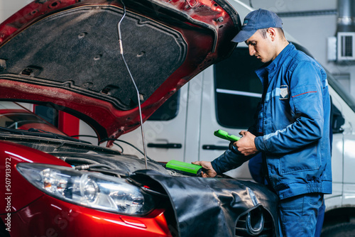 Car service electrician or mechanic uses a tablet computer with futuristic interactive diagnostics software. Inspecting the vehicle in order to find broken components in the engine bay of modern car