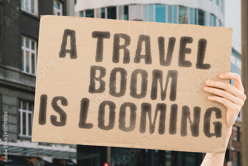 The phrase " A travel boom is looming " is on a banner in men's hands with blurred background. Gain. Burst. Explosion. Explode. Rise. Increase. Progress. Flying. Global. Rising. Rate. Profit. Improve © AndriiKoval