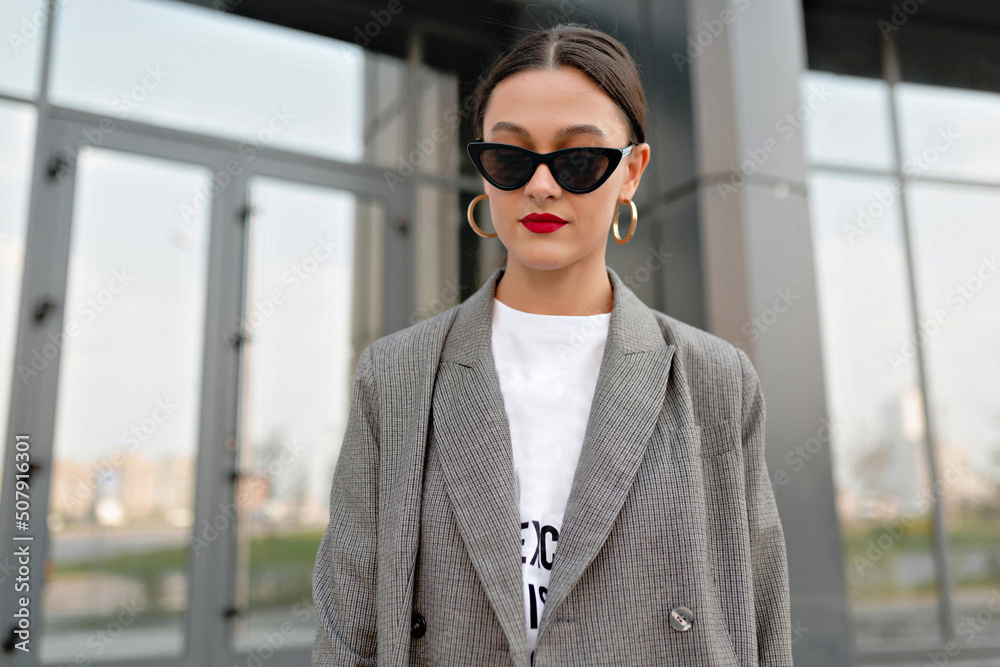 Incredible stylish woman with dark hair and red lips wearing golden earrings and grey jacket posing at camera outdoor against glass building