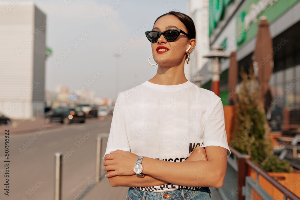 Outside portrait of elegant stylish woman with dark hair wearing sunglasses and white t-shirt posing in city in sunlight. Stylish woman in spectaculars standing outdoors