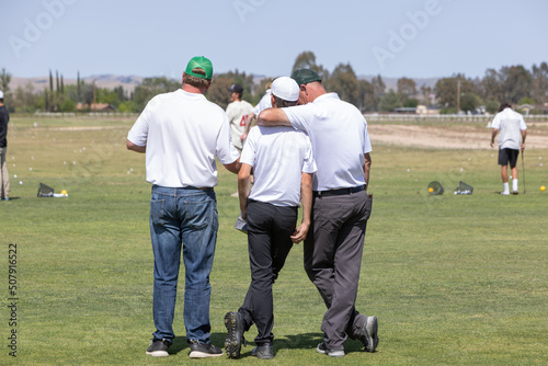 Looking at the back of three men standing on the golf course. One man talking to another with his arm around his shoulder. Golf coach or father giving instruction.