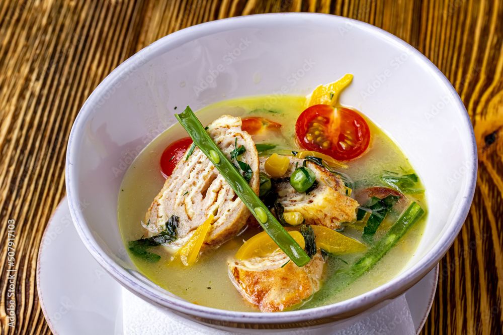 Vegetable soup with chicken rolls is a soup with the main ingredients of carrots, broccoli, celery and green onions, and sliced chicken rolls. Served in a black bowl on the table. Selective focus