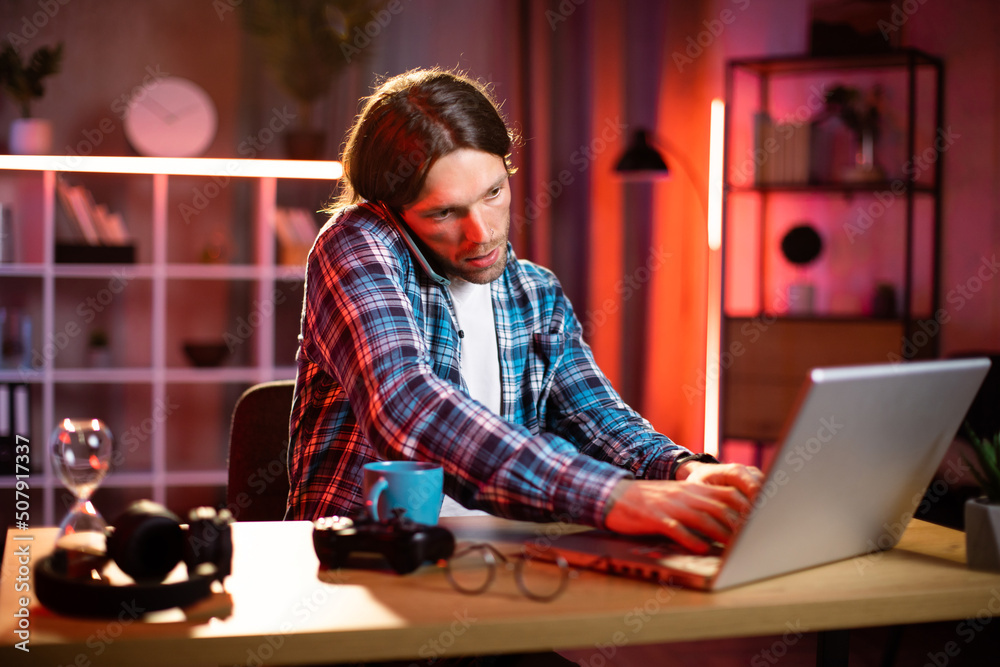 Attactive satisfied smiling young caucasian man in checkered shirt talking on phone and typing on laptop, while sitting at the table in modern decorated living room at home . Online work concept