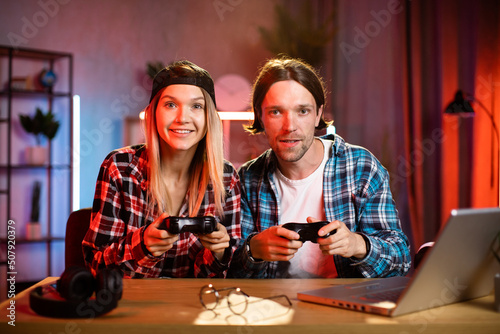 Young multiracial couple sitting at table with wireless joysticks in hands and playing video games on laptop. Caucasian man and blond woman in black cap spending evening time at home with fun.