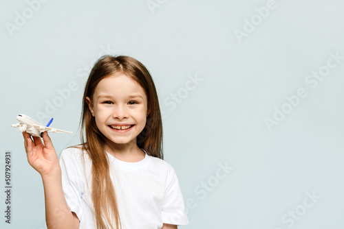 Little girl smiling while holding a fake airplane on a blue background and looking at the camera. Tourism and travel concept. Copy space