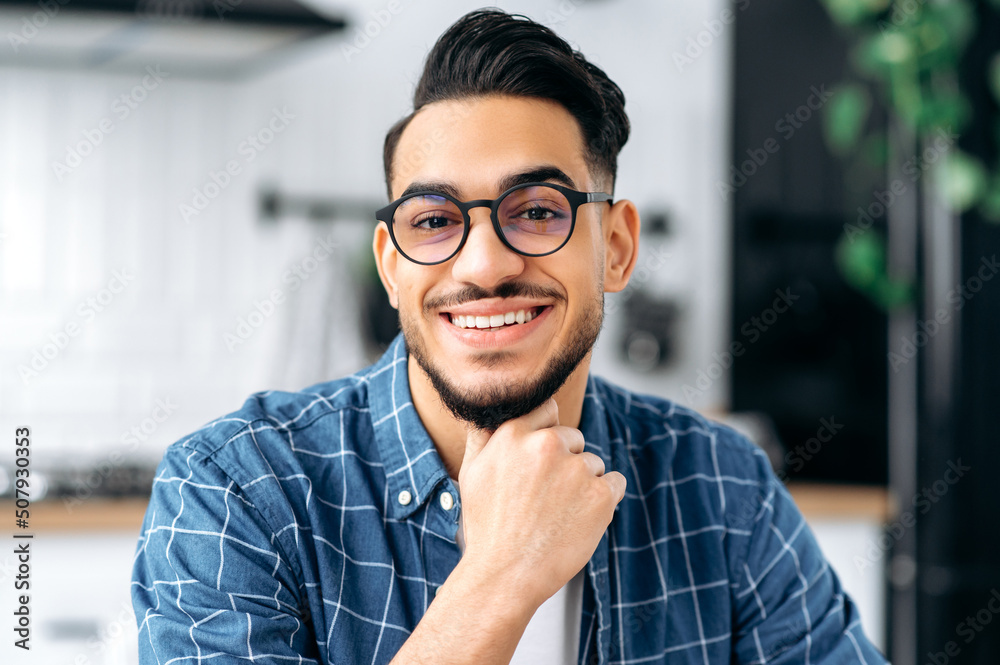 Obraz premium Close-up portrait of attractive positive arabian or indian young man with glasses, freelancer or student, in casual clothes, with perfect white teeth, looking at the camera with toothy happy smile