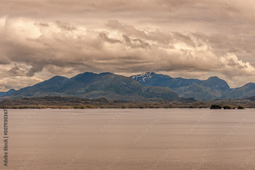 Pacific Coastline, Alaska, USA - July 16, 2011: Canadian Rocky mountains under heavy brown cloudscape with few green forested islets along brown water coastline. Patches of snow. Pacific Coastline, Alaska, USA - July 16, 2011: Canadian Rocky mountains under heavy brown cloudscape with few green forested islets along brown water coastline. Patches of snow.