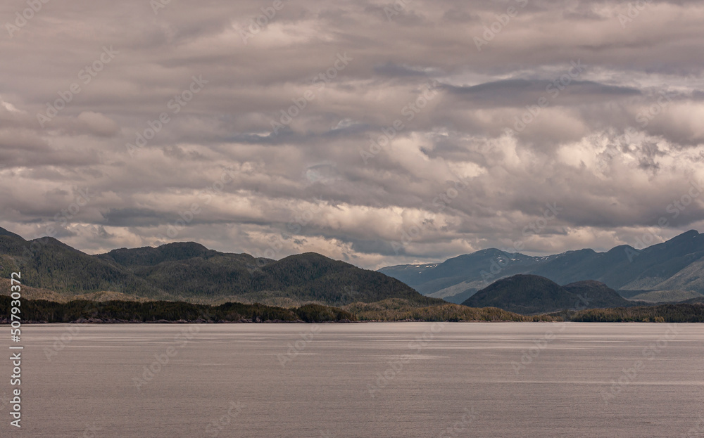 Pacific Coastline, Alaska, USA - July 16, 2011: Canadian Rocky mountains under heavy gray-white cloudscape along thick green forested hilly coastline behind brownish flat Pacific Ocean water.