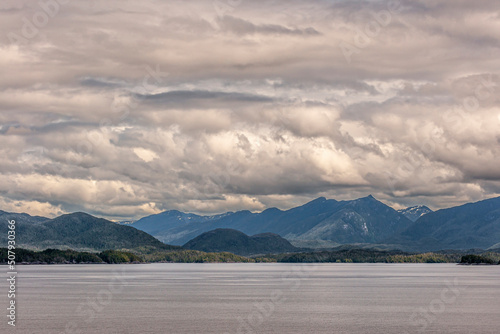 Pacific Coastline, Alaska, USA - July 16, 2011: Canadian Rocky tall mountains under heavy white and gray cloudscape along thick green forested hilly coastline behind brownish flat Pacific Ocean water. © Klodien