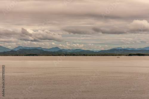 Pacific Coastline, Alaska, USA - July 16, 2011: Canadian Rocky mountains under heavy cloudscape with green forested coastline. Small fishing vessel om brown Pacific water.