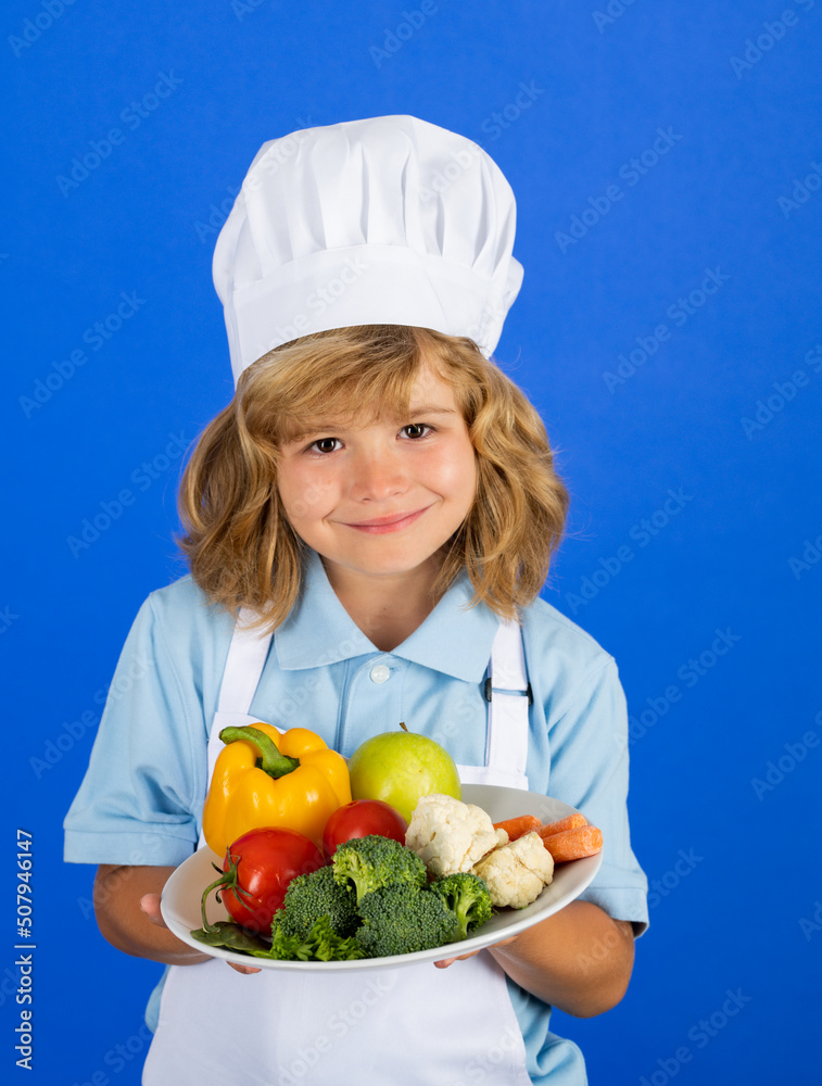 Kid boy in chef hat and apron hold plate with vegetables cooking preparing meal. Little cook with vegetables at kitchen. Natural kids food. Kid boy in chef hat and apron hold plate with vegetables cooking preparing meal. Little cook with vegetables at kitchen. Natural kids food.