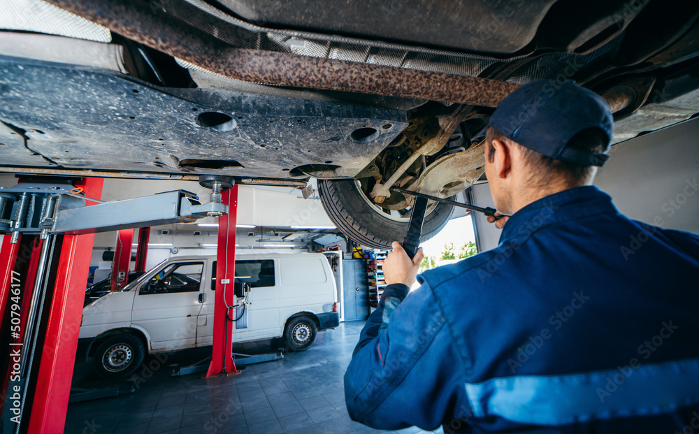 Fototapeta premium Young car mechanic at repair service station inspecting car wheel and suspension detail of lifted automobile. Bottom view.