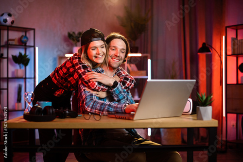 Focused caucasian man working on modern laptop during evening time and his girlfriend hugs him from back. Concept of people, home routine, freelance and deadline.