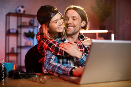 Focused caucasian man working on modern laptop during evening time and his girlfriend hugs him from back. Concept of people, home routine, freelance and deadline.