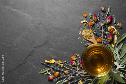 Glass cup of aromatic freshly brewed tea near different dry herbs on black table, flat lay. Space for text