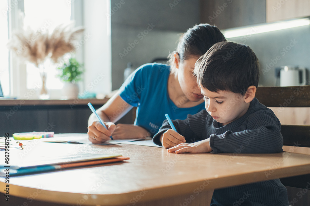 Mother and son writing in notepad while sitting together at table and helping son to do homework. Homework assignment. Adult woman teaches the child the Mother and son writing in notepad while sitting together at table and helping son to do homework. Homework assignment. Adult woman teaches the child the