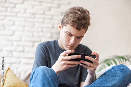 A teenage boy plays games on a smartphone with a focused expression. Dependence on the phone and online games. Close-up, bottom view. A young man with a phone in his hands sits on the couch.