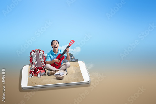 Asian man with a beanie hat and a backpack playing guitar to make money on the beach © Leo Lintang