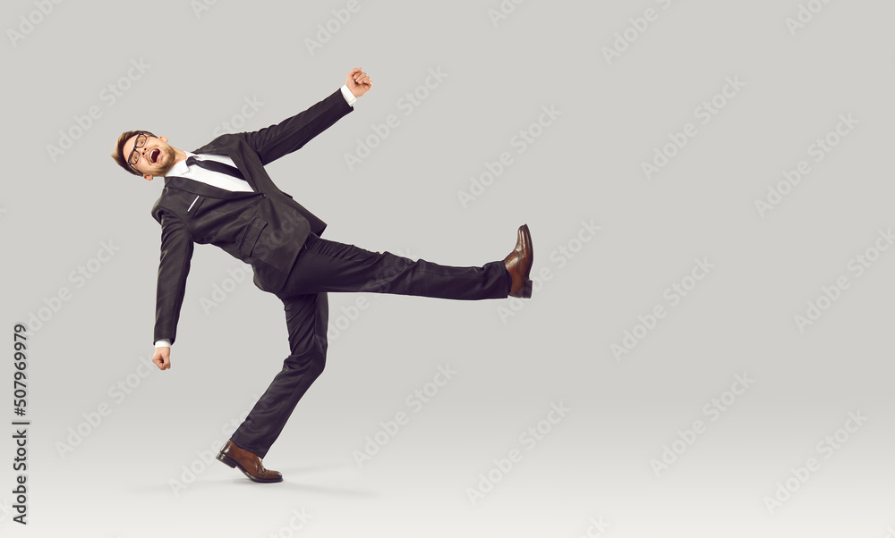 Businessman or office worker behaving in very strange way. Full body length portrait of funny young business man in suit with crazy weird face expression walking against light gray studio background