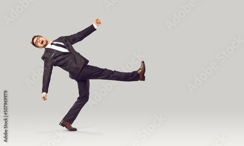 Businessman or office worker behaving in very strange way. Full body length portrait of funny young business man in suit with crazy weird face expression walking against light gray studio background