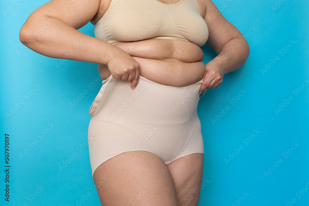 Woman with folds on stomach pulling beige shapewear panties over belly  closeup. Tighten figure and wear lifting underwear for weight loss, blue  background. Concept of diet and fighting overweight. Photos