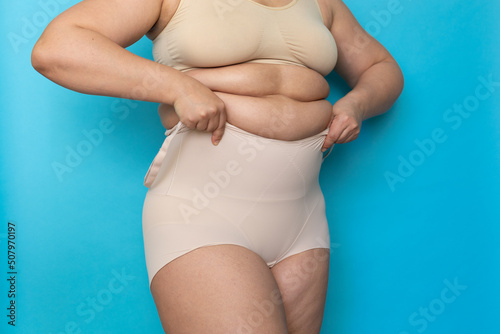 Woman with folds on stomach pulling beige shapewear panties over belly closeup. Tighten figure and wear lifting underwear for weight loss, blue background. Concept of diet and fighting overweight. © Юля Бурмистрова