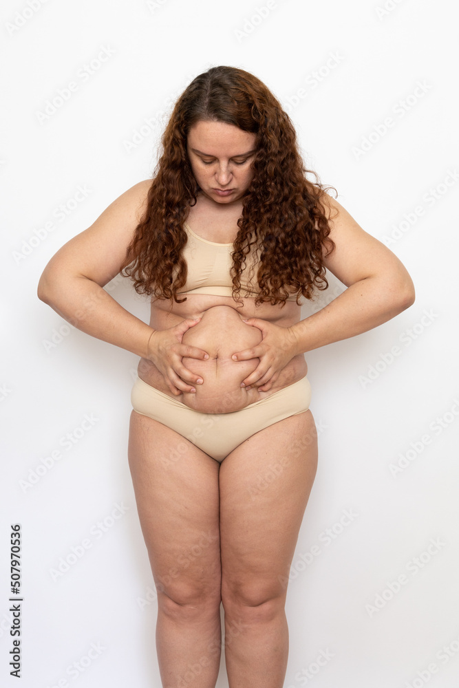 Portrait worried plump woman with curly hair wearing beige underwear, squeezing excess fat of belly, bowing head, looking at stomach on white background. Weight loss, obesity, diet, health problems. Portrait worried plump woman with curly hair wearing beige underwear, squeezing excess fat of belly, bowing head, looking at stomach on white background. Weight loss, obesity, diet, health problems.