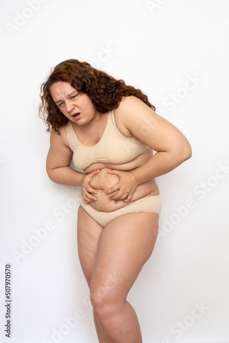 Portrait of upset plump woman with curly hair wearing beige underwear, squeezing excess subcutaneous fat of belly, having abdominal pain on white background. Weight loss, stomachache, health problems.