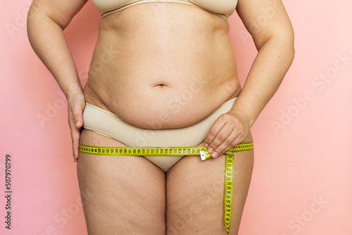Cropped photo of naked overweight woman body, holding and measuring by roulette tape her hips, buttocks. Large and big size fat tummy. Folds on her stomach. Unhealthy, obesity problem. Disproportion