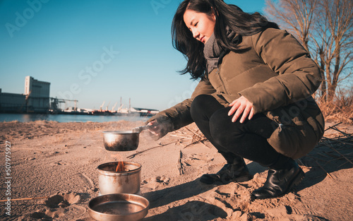 Young asian woman with warm clothes on is cooking with outdoor cooker on Harriersand beach at a riverbank during golden hour. Weser river blurred in the background.  photo