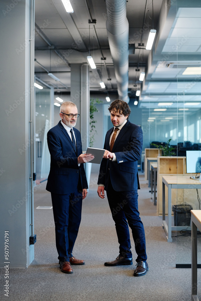 Two businessmen in formal wear discussing online presentation on digital tablet while standing at office Two businessmen in formal wear discussing online presentation on digital tablet while standing at office