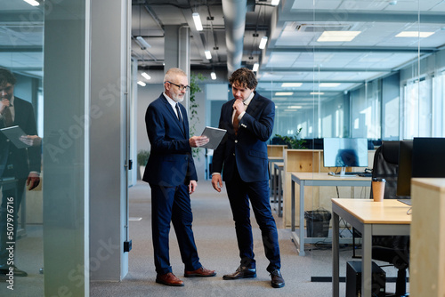 Mature businessman in suit showing something on digital tablet to his colleague while they standing at office
