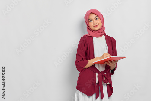 Pensive beautiful Asian woman in casual shirt holding notebook and thinking about something isolated over white background