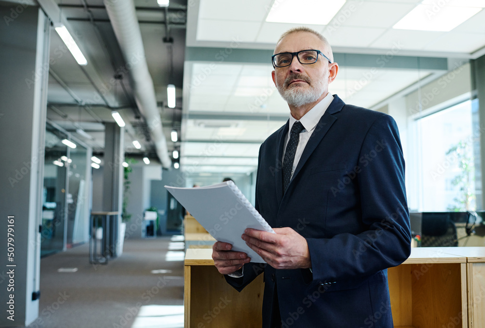Fototapeta premium Portrait of senior businessman in dark suit holding report and looking at camera while standing at office hall