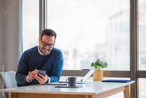 Smiling businessman socializing on smart phone while working at desk in office