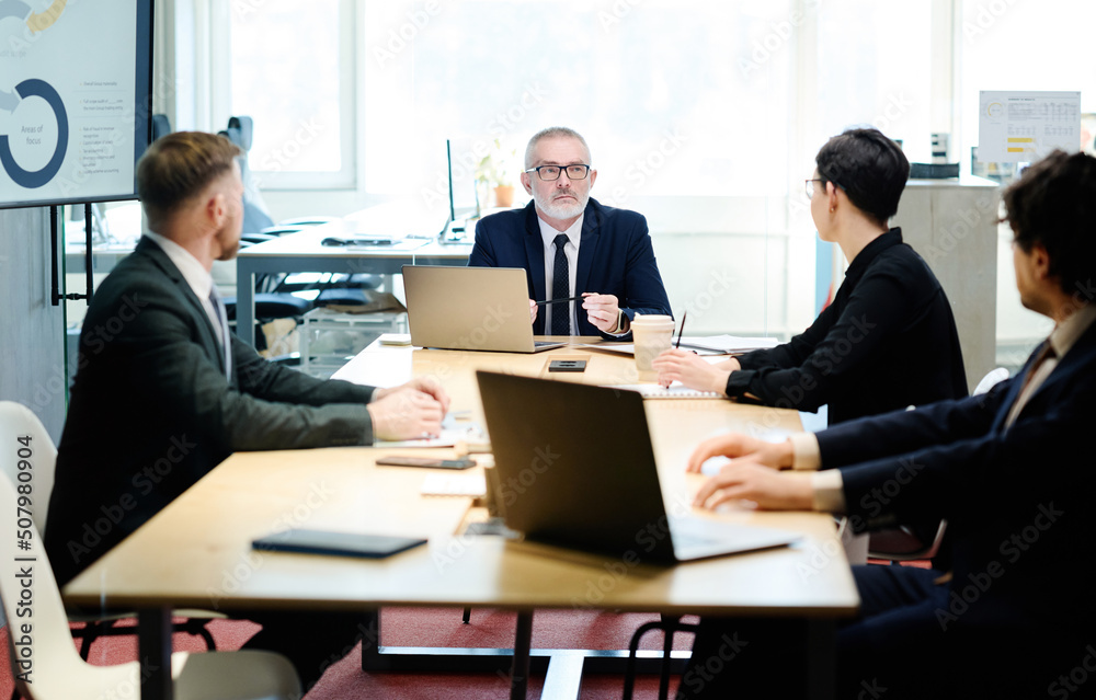 Senior manager in suit talking to partners at table with laptops during meeting at meeting room Senior manager in suit talking to partners at table with laptops during meeting at meeting room
