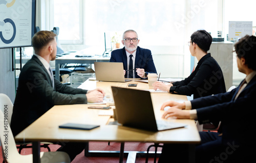 Senior manager in suit talking to partners at table with laptops during meeting at meeting room