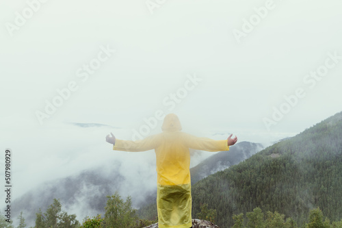 A man in a yellow raincoat stands on top of a mountain on a rock and looks at a beautiful misty view with his hands raised.